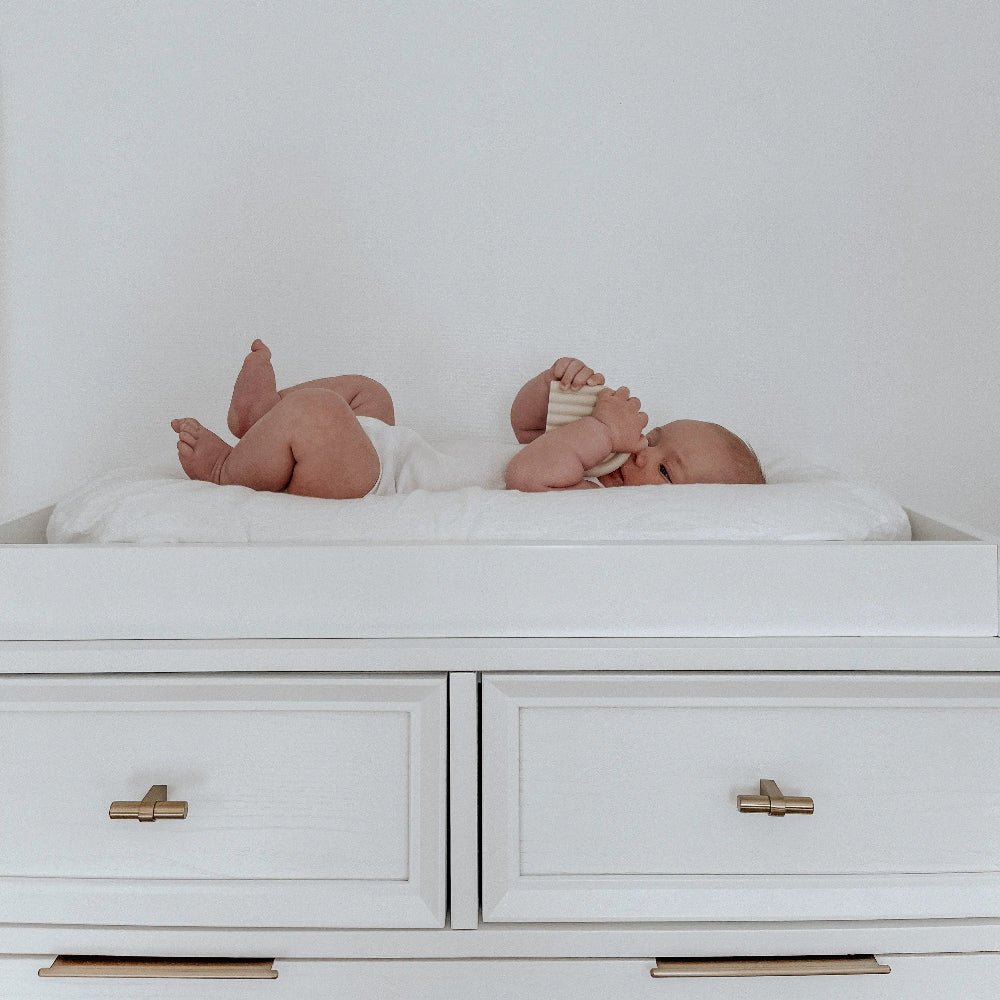 A baby lying on a white changing pad placed on top of a white dresser with drawers, featuring a simple design with metal handles.